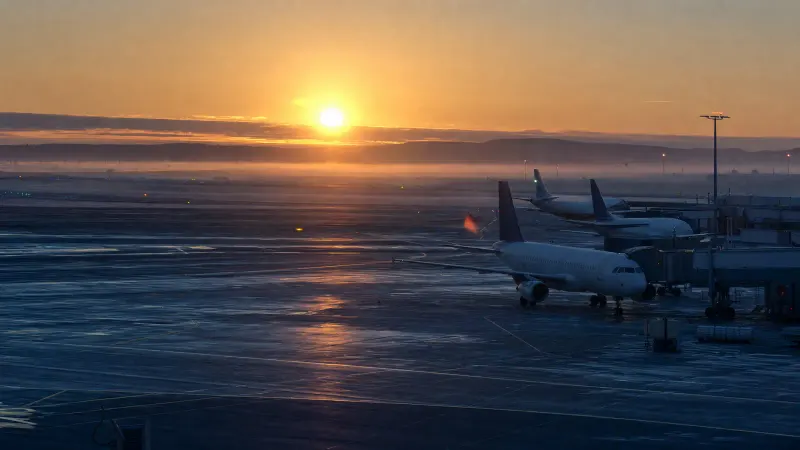 Parked passenger jets on a calm airport apron at dawn under a soft golden sky with light mist over the tarmac.